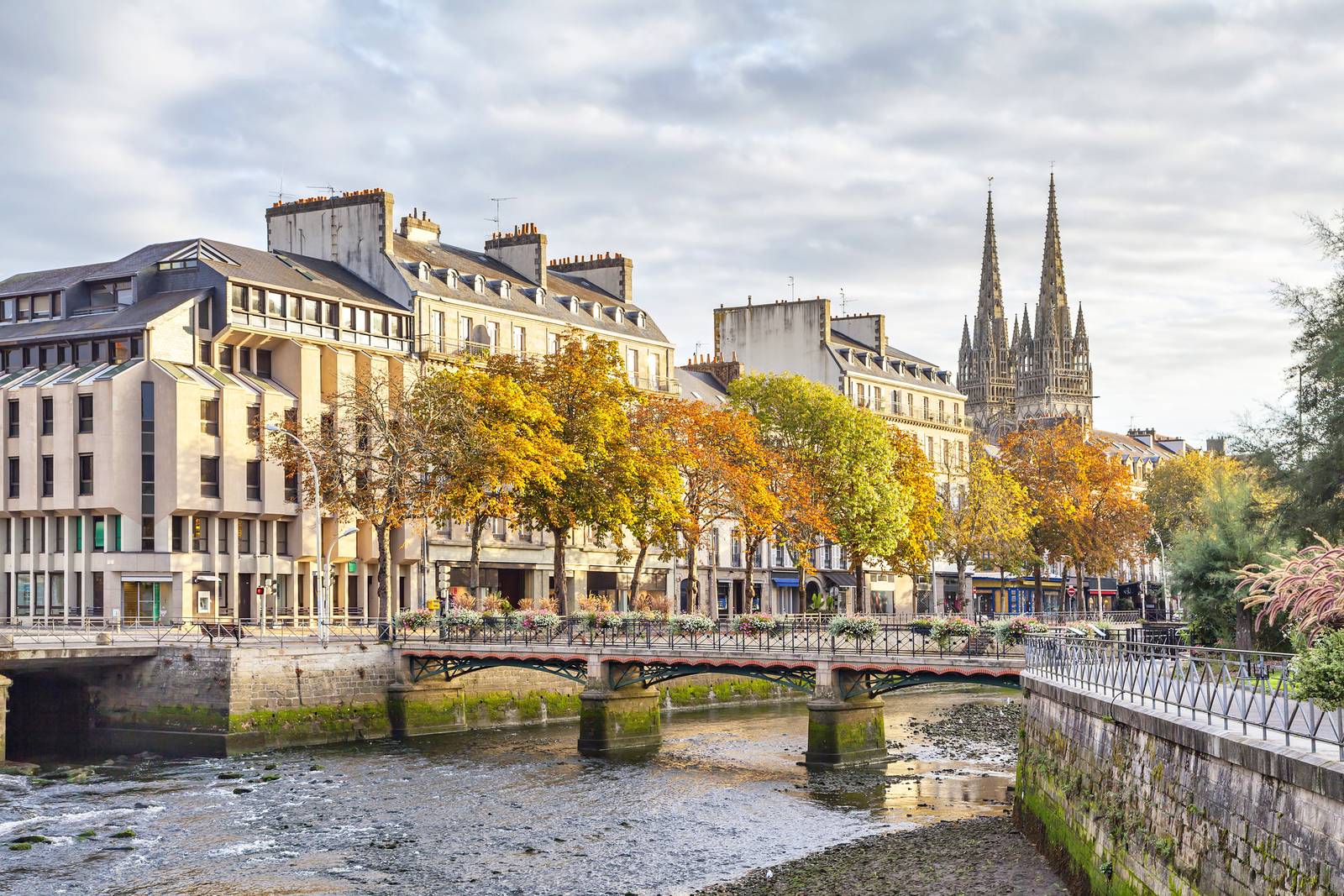 A scenic view of Quimper with historic buildings, a river, a small bridge, and trees with autumn foliage under a cloudy sky.