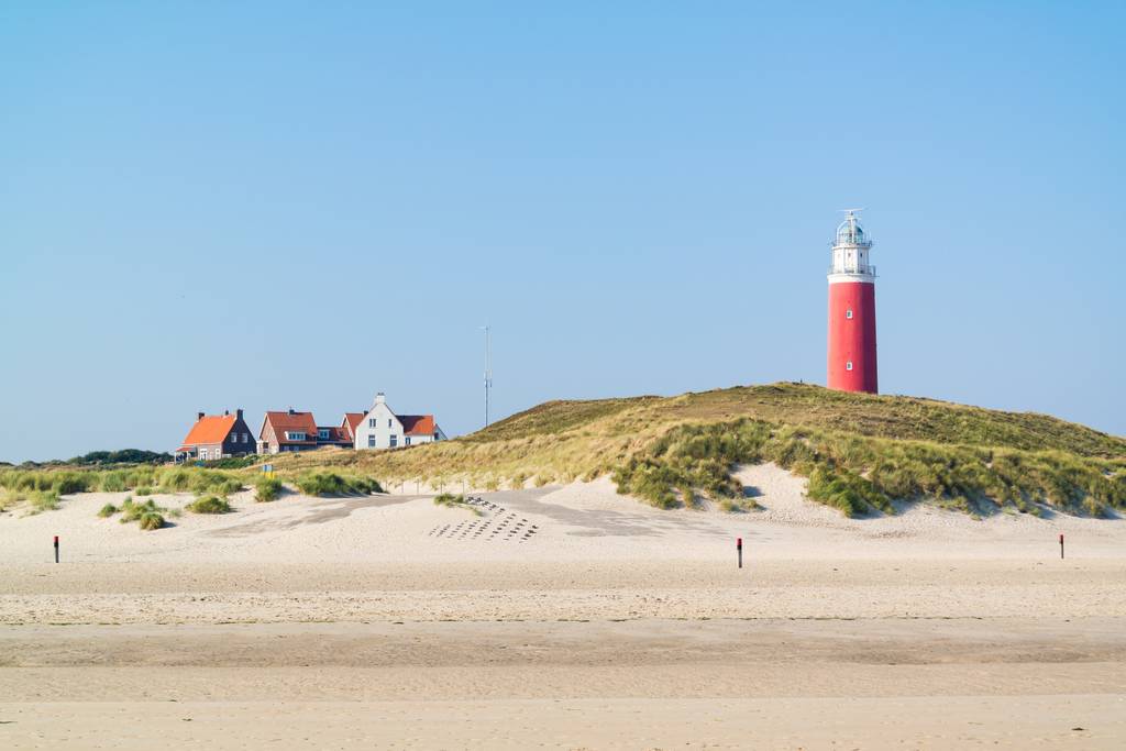 Roter Leuchtturm auf grasbewachsenen Sanddünen neben weißen Häusern mit orangen Dächern, breiter Sandstrand und klarer blauer Himmel auf Texel..