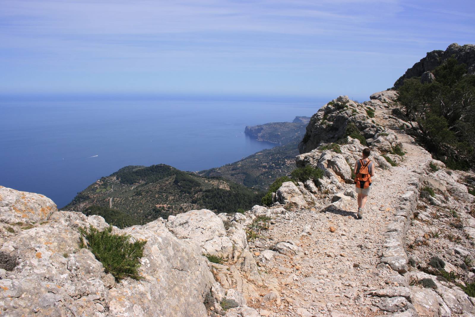 Wanderer mit orangefarbenem Rucksack auf einem felsigen Bergpfad entlang eines Küstenkamms mit Blick auf das blaue Meer und entfernte Klippen.
