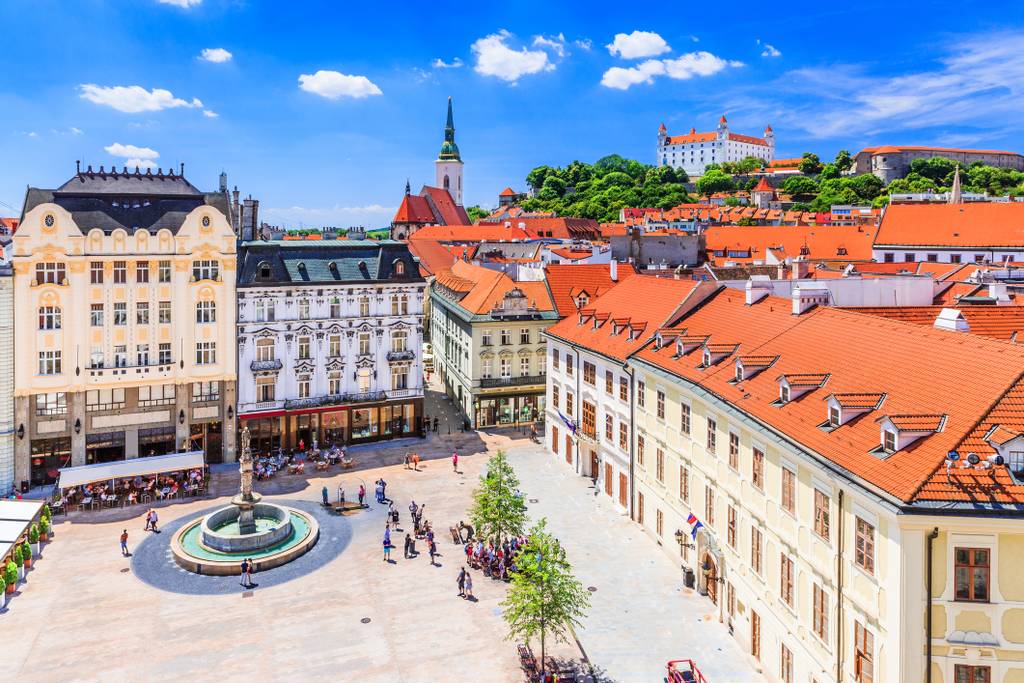 Aerial view of a European city square with historic buildings, a fountain, and a castle on a hill under a blue sky.