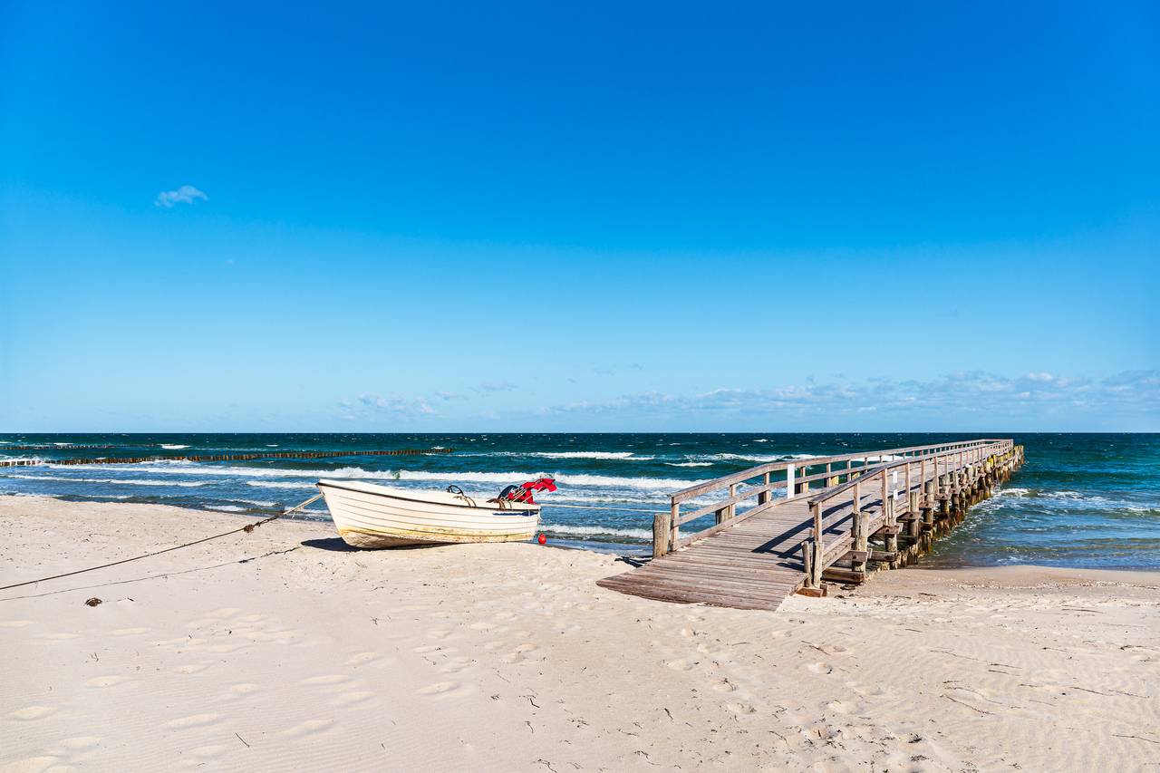 Ein Holzboot liegt an einem Sandstrand in der Nähe eines hölzernen Stegs, der in das ruhige Meer unter einem klaren blauen Himmel ragt.