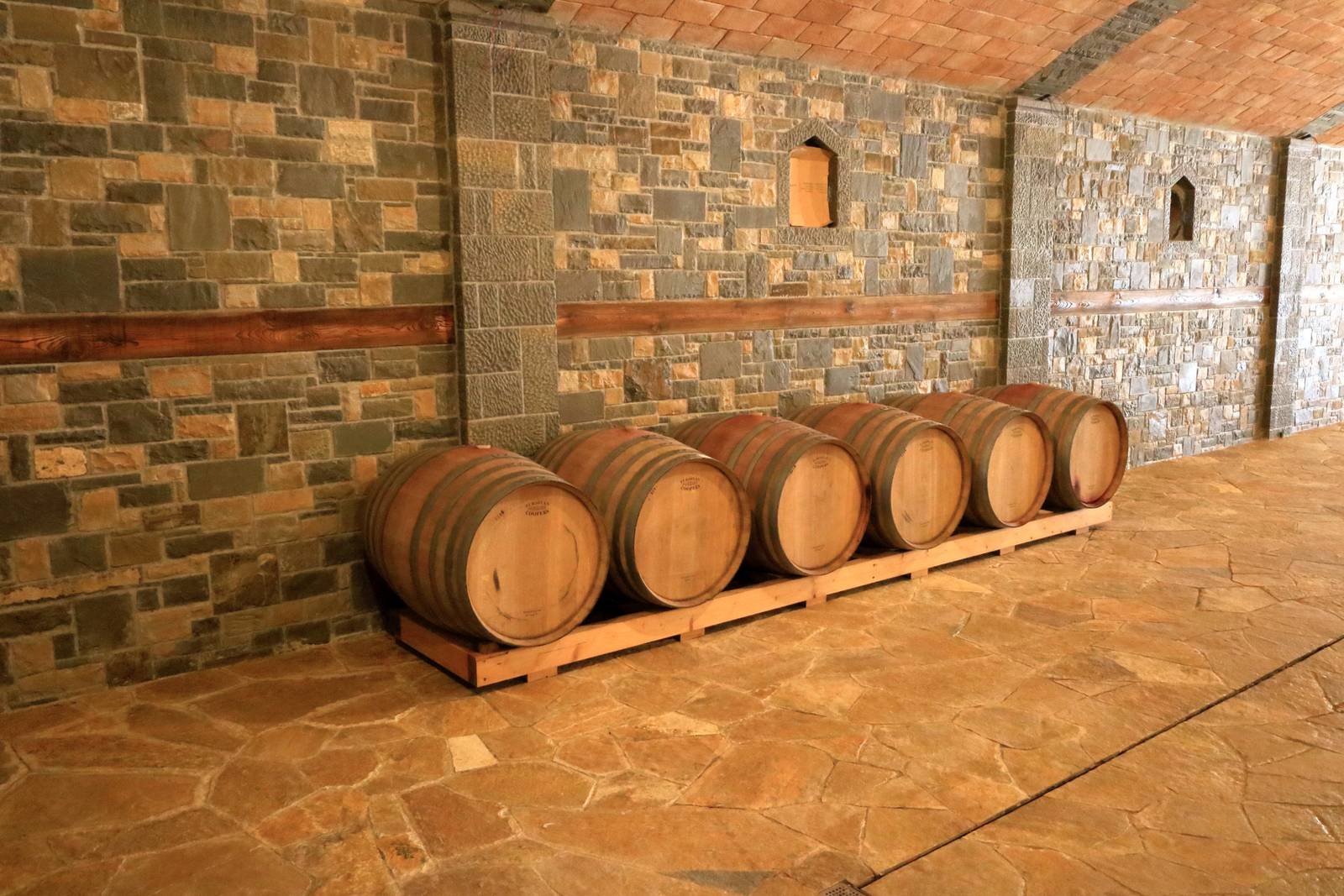 Fünf Holzfässer auf einem Holzgestell vor einer Steinwand in einem großzügigen Raum mit gefliestem Boden und Decke