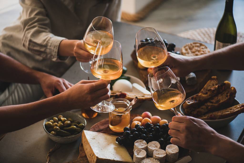 Vier Personen stoßen mit Gläsern Weißwein über einem Tisch mit Käse, Trauben, Oliven und Brot an – gemütliche und einladende Atmosphäre