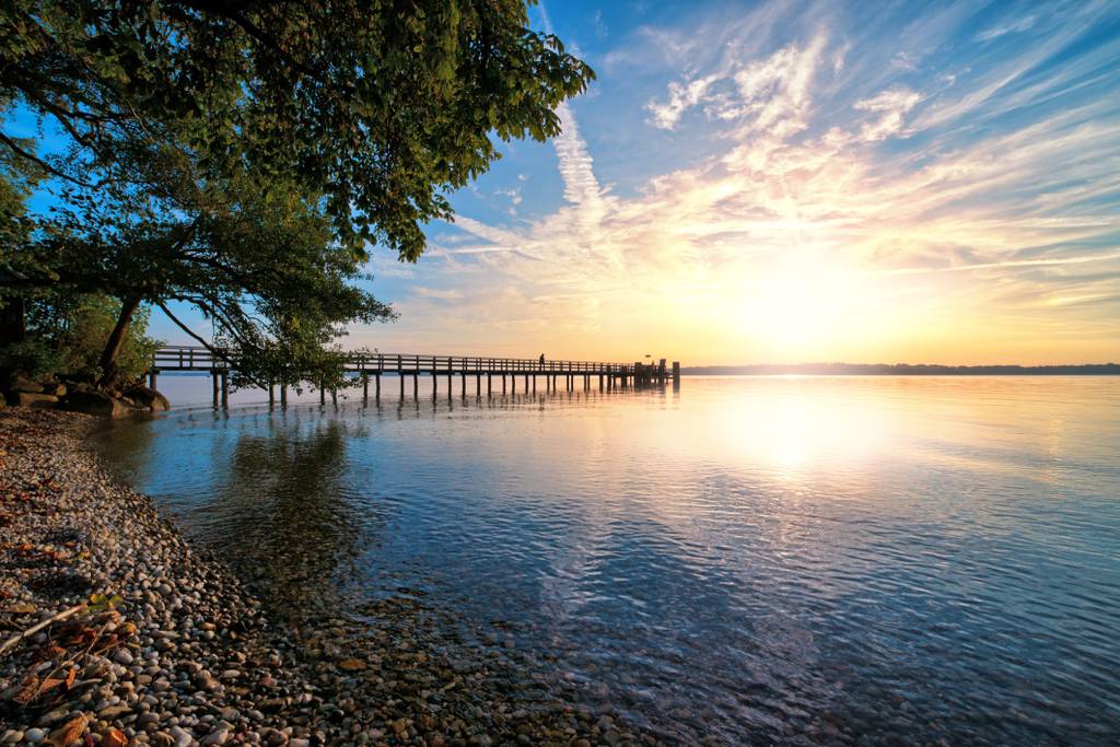 Sonnenuntergang über einem ruhigen See mit einem langen Holzsteg, üppigen Bäumen am Ufer und einem bunten Himmel, der sich im stillen Wasser spiegelt.