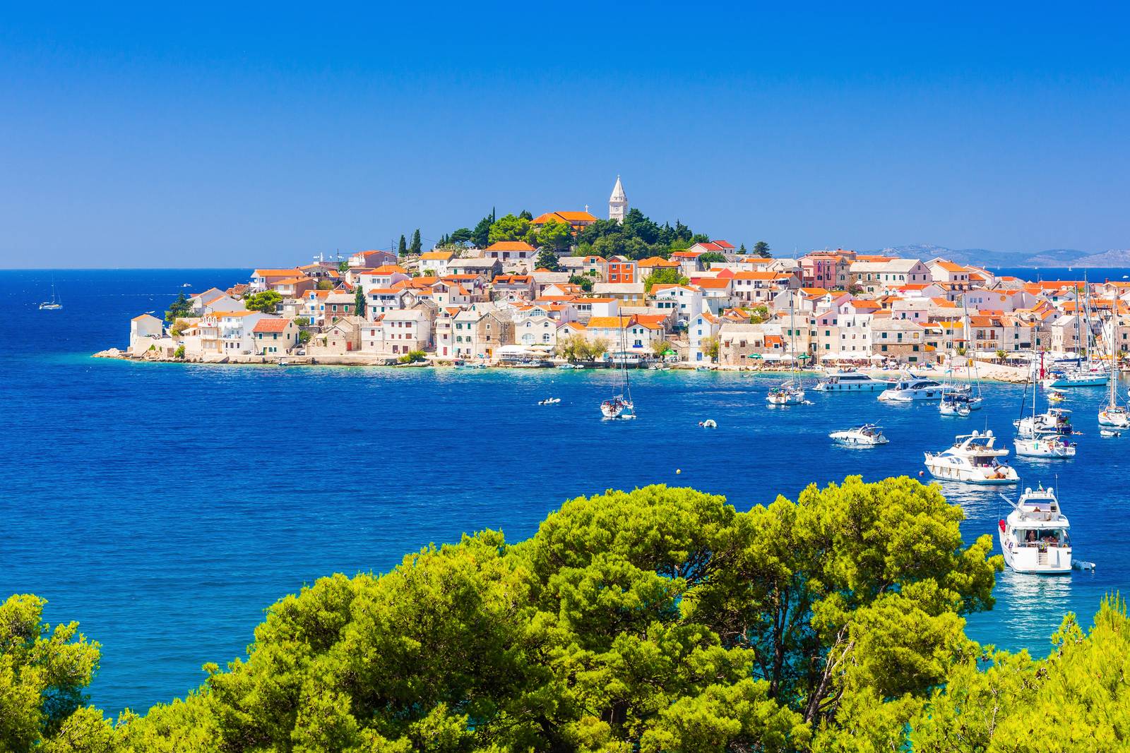 Primosten view with orange-roofed buildings, surrounded by blue sea and boats, with lush greenery in the foreground under a clear blue sky.