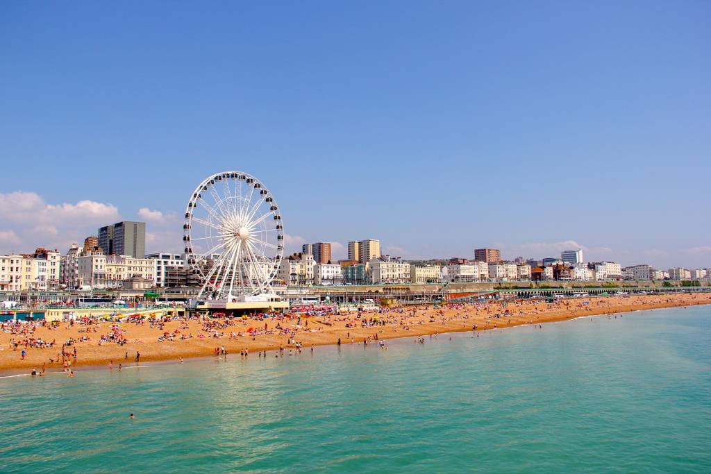 Beachfront cityscape of Brighton with a Ferris wheel, crowded sandy beach, and calm turquoise sea under a clear blue sky.