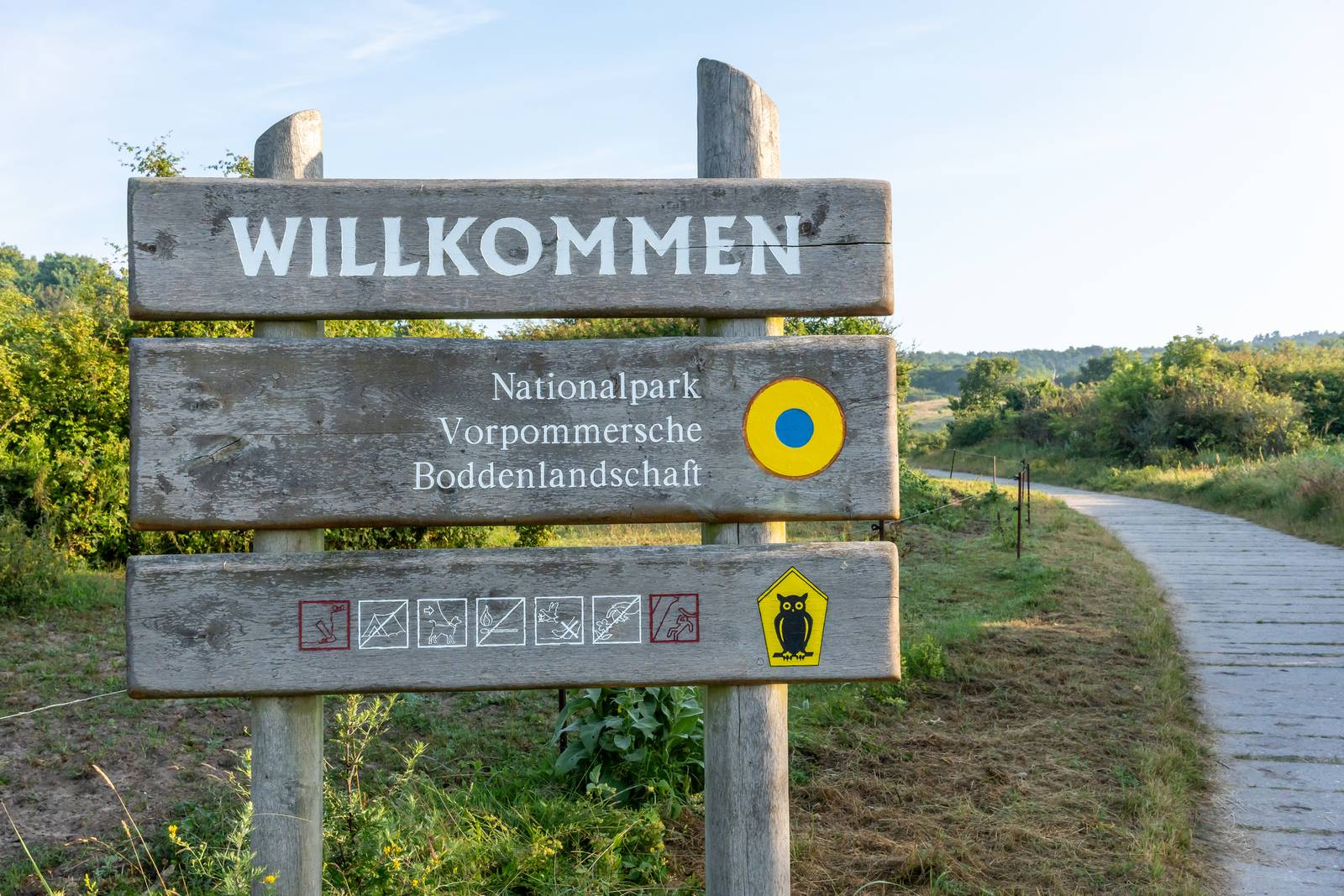 Holzschild mit der Aufschrift „Willkommen“ am Eingang des Nationalparks Vorpommersche Boddenlandschaft, im Hintergrund ein Naturpfad und Grünflächen.