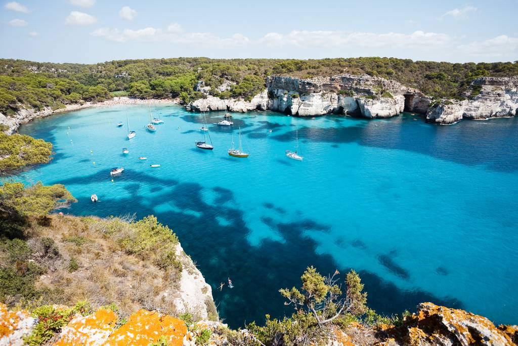 Vista panoramica di Cala Macarella, una baia turchese con barche ancorate, circondata da scogliere rocciose e vegetazione lussureggiante sotto un cielo azzurro con alcune nuvole.