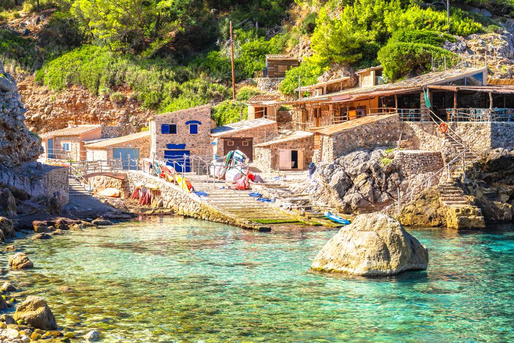 Scenario costiero vicino a Cala Deià con edifici in pietra rustica, acqua turchese cristallina, rocce e vegetazione abbondante in una giornata soleggiata.
