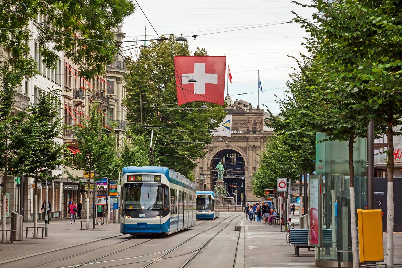 Straßenbahnen auf einer baumbestandenen Straße in Zürich, Schweiz, mit einer Schweizer Flagge darüber und einem historischen Torbogen im Hintergrund.
