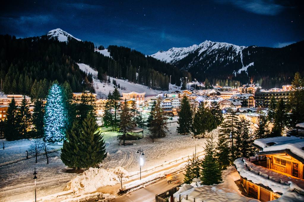 Verschneites Alpendorf bei Nacht mit beleuchteten Gebäuden, einem festlich geschmückten Weihnachtsbaum und einer Bergkulisse unter einem sternenklaren Himmel.