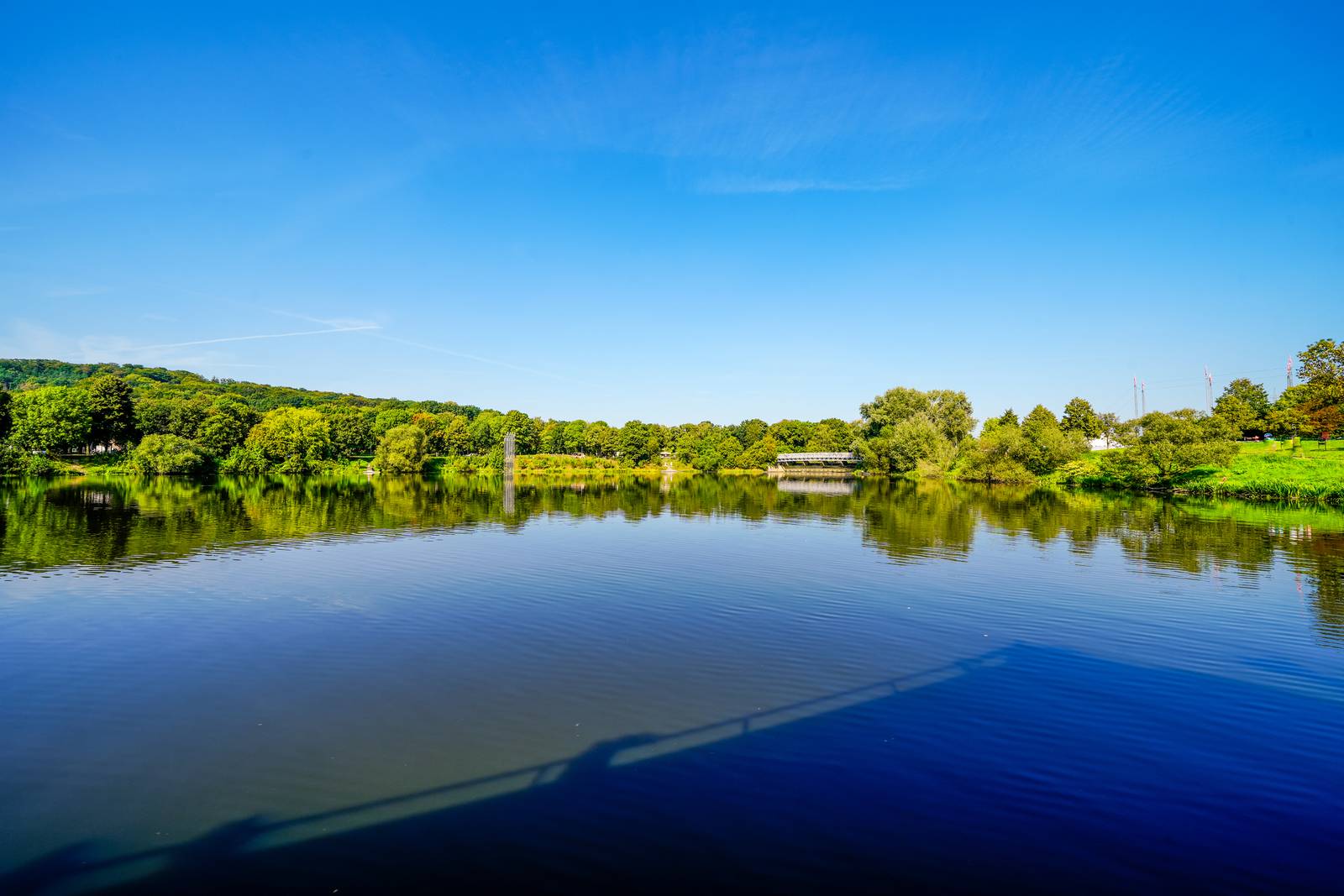 Ein ruhiger See, der einen klaren blauen Himmel spiegelt, umgeben von üppig grünen Bäumen und einer entfernten Brücke unter hellem Sonnenlicht