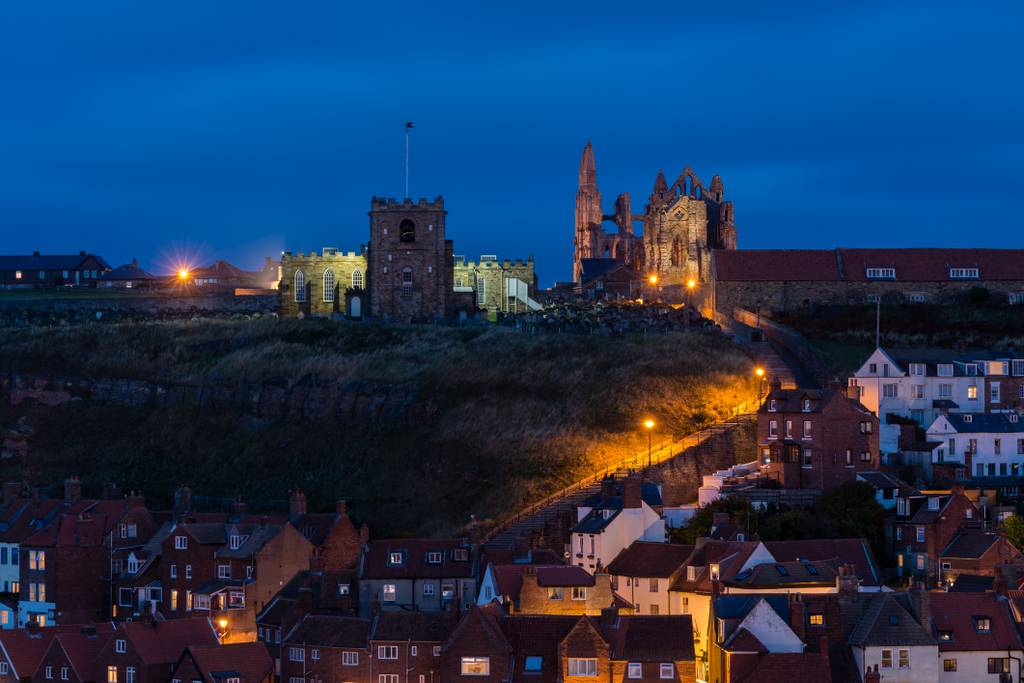 Night view of Whitby Abbey and St. Mary's Church on a hill, illuminated amidst a seaside town with cozy lights and a cloudy blue sky.