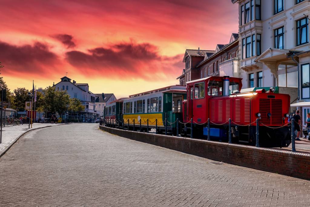 Roter Touristenzug mit grünen und gelben Waggons, der entlang einer kopfsteingepflasterten Straße neben Gebäuden am Wasser unter einem dramatischen orange-rosafarbenen Sonnenuntergang steht.