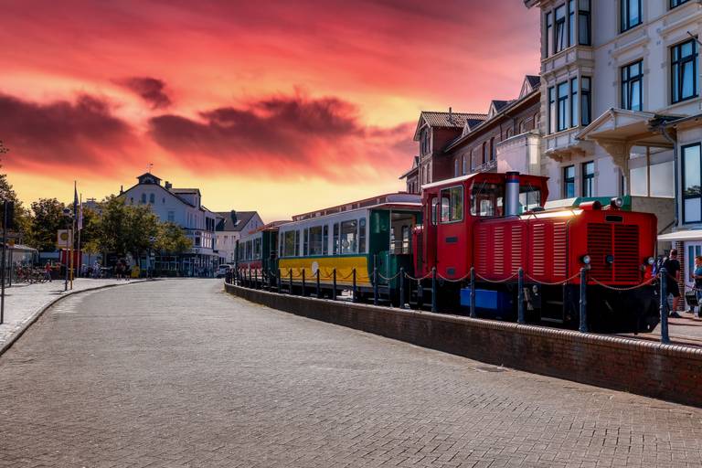 Roter Touristenzug mit grünen und gelben Waggons, der entlang einer kopfsteingepflasterten Straße neben Gebäuden am Wasser unter einem dramatischen orange-rosafarbenen Sonnenuntergang steht.