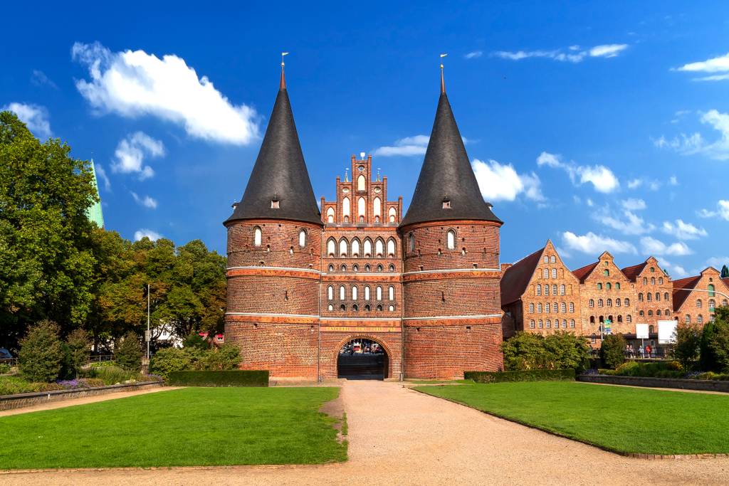 Holstentor, brick Gothic city gate in Lübeck with twin conical towers, central archway, grassy forecourt and path under a bright blue sky.
