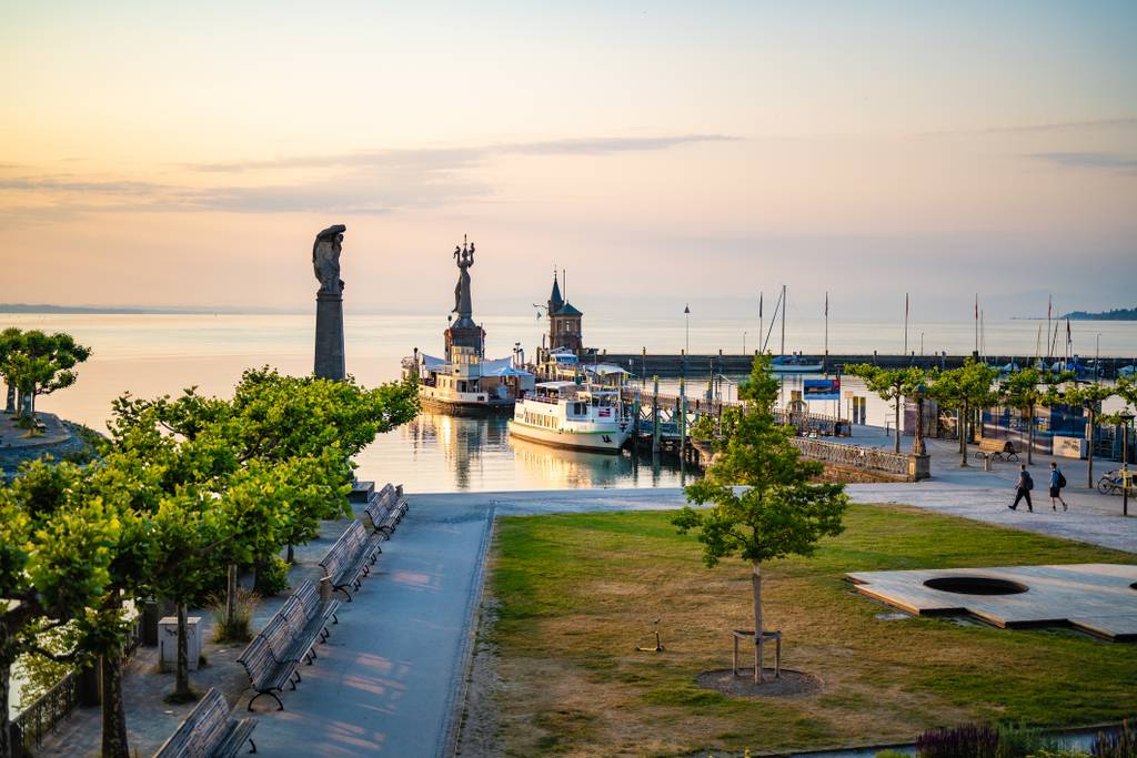 Hafen bei Sonnenuntergang mit festgemachten Booten, zwei Skulpturen, von Bäumen gesäumten Wegen und ruhigem Wasser, das die warmen Farbtöne des Himmels widerspiegelt.