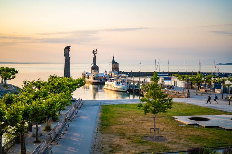 Hafen bei Sonnenuntergang mit festgemachten Booten, zwei Skulpturen, von Bäumen gesäumten Wegen und ruhigem Wasser, das die warmen Farbtöne des Himmels widerspiegelt.