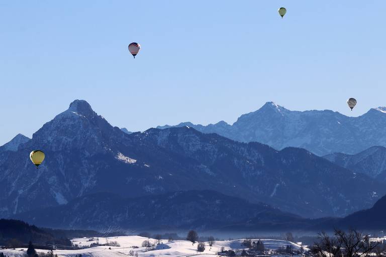 Fünf bunte Heißluftballons treiben über schneebedeckte Felder auf die gestuften blauen Alpenberge zu, unter klarem Himmel.