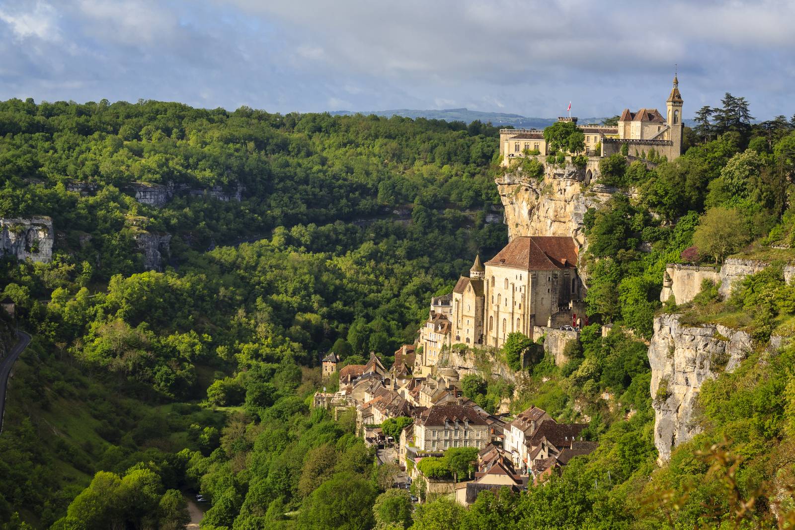 Medieval village of Rocamadour, France, perched on a cliffside surrounded by lush green forests under a partly cloudy sky.