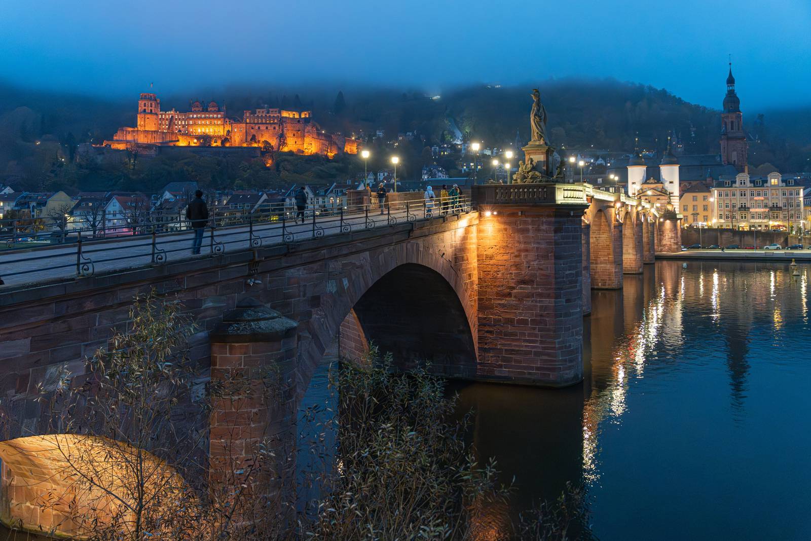 Beleuchtete Brücke über einem ruhigen Fluss in der Dämmerung, im Hintergrund ein erleuchtetes Schloss auf einem Hügel, umgeben von Nebel und Stadtlichtern.