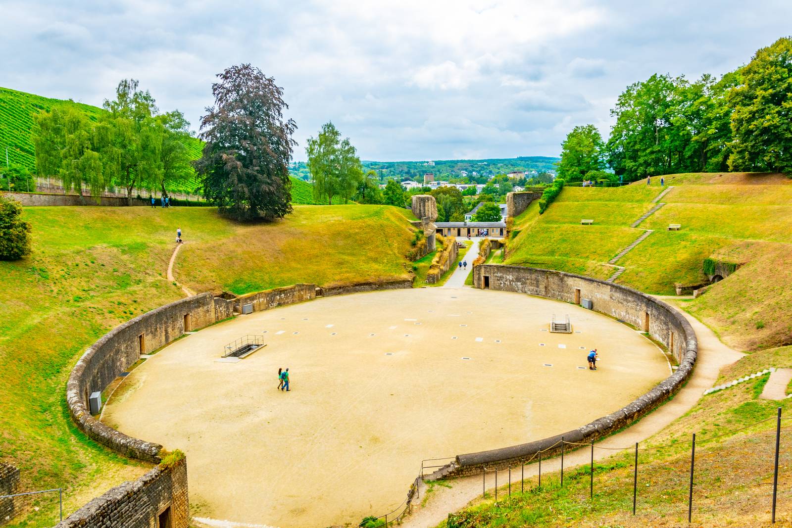 Antikes Amphitheater mit Steinsitzreihen, umgeben von grünen Hügeln und Bäumen unter einem wolkigen Himmel, mit einigen Menschen, die im Inneren umhergehenAncient amphitheater with stone seating, surrounded by green hills and trees under a cloudy sky, with a few people walking inside.