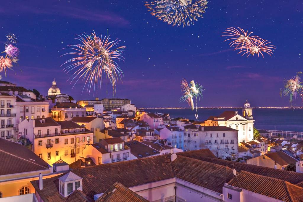 Fireworks illuminate the night sky over a historic coastal city with colorful buildings and a view of the ocean.