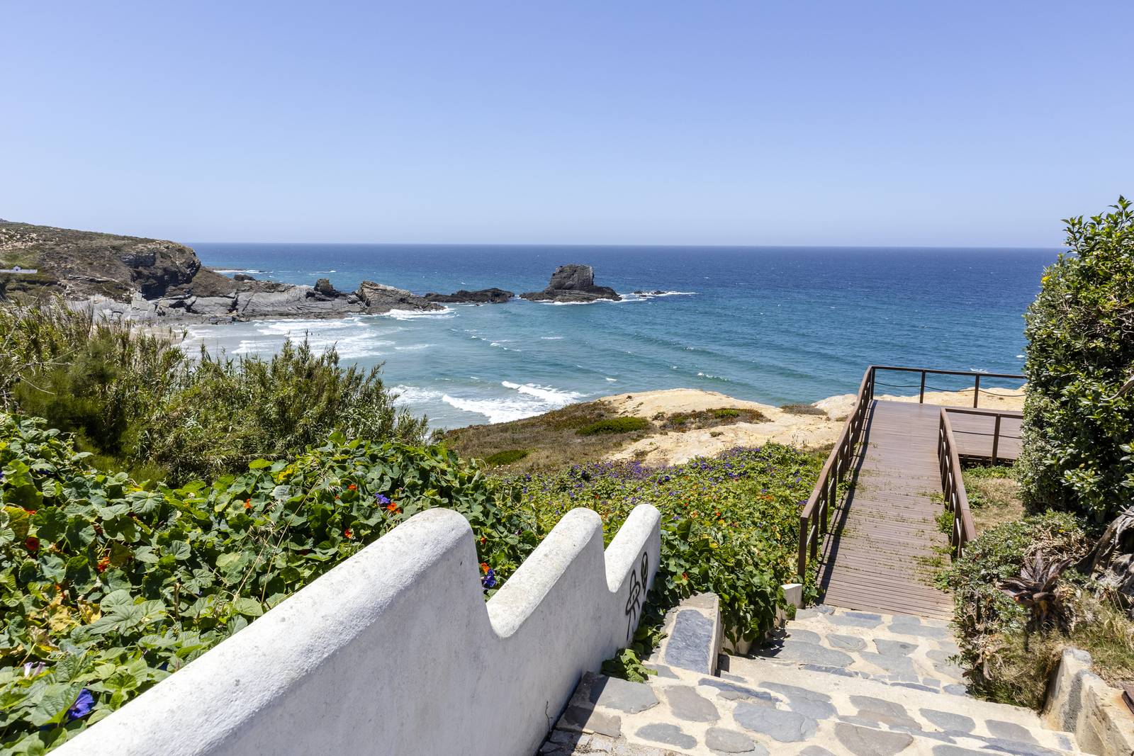 Stone steps with white railing lead to a wooden path overlooking a sandy beach and blue ocean under a clear sky.