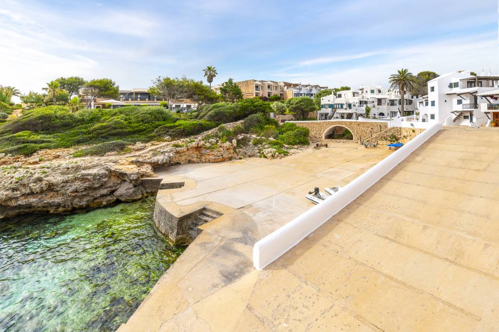 Vista costiera a Binibeca Vell  con acqua turchese cristallina, costa rocosa ed edifici bianchi circondati da vegetazione sotto un cielo azzurro.