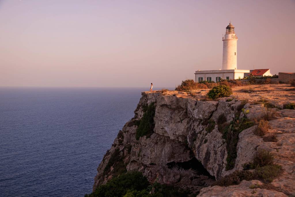 Faro situato su una scogliera rocciosa con vista su un oceano immenso al tramonto, con una persona vicino al bordo che contempla l'orizzonte.