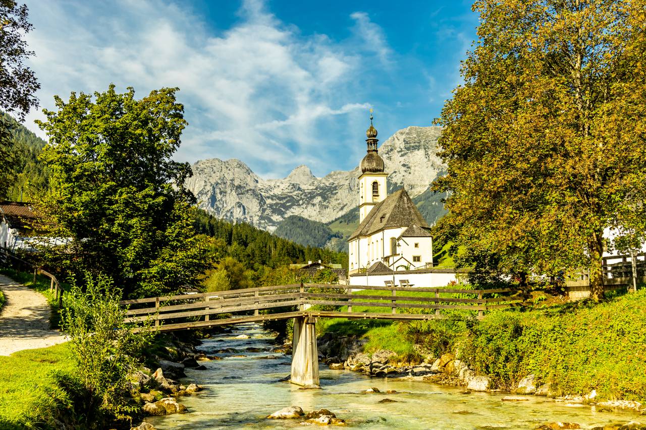 Idyllische Ansicht einer rustikalen Kirche neben einer Holzbrücke über einem klaren Bach, mit majestätischen Bergen und üppigen Bäumen unter einem blauen Himmel.