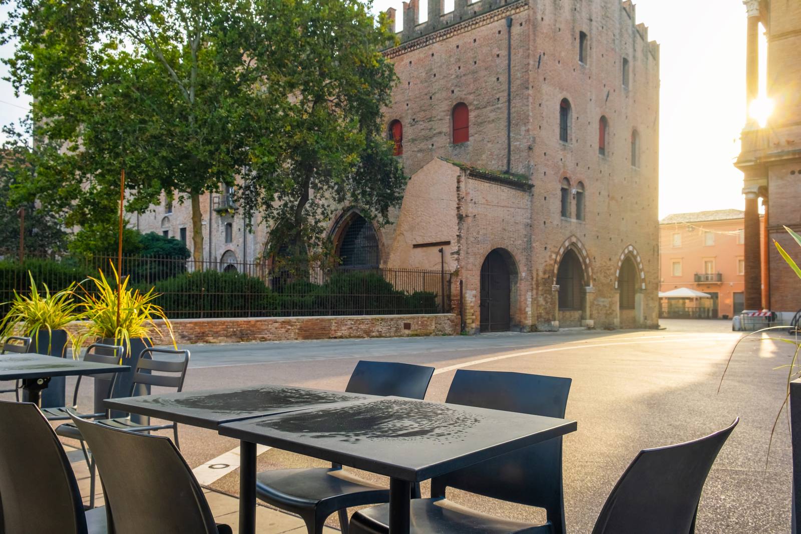 Straßencafé mit leeren Tischen und Stühlen, gegenüber einem historischen Backsteingebäude und einem Baum, im warmen Sonnenlicht. Ruhige Straße im Hintergrund