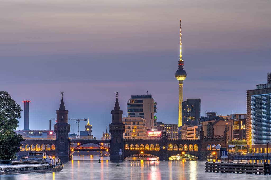 Dämmerungsblick auf die Oberbaumbrücke über die Spree, beleuchtete Bögen mit Spiegelungen, im Hintergrund ragt der Berliner Fernsehturm über die Stadtsilhouette.