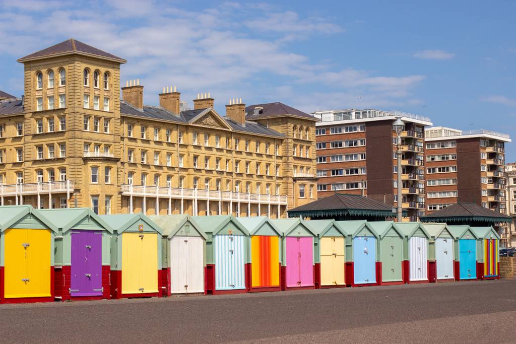 Colorful beach huts line a promenade with a historic hotel and modern apartments in the background under a blue sky.