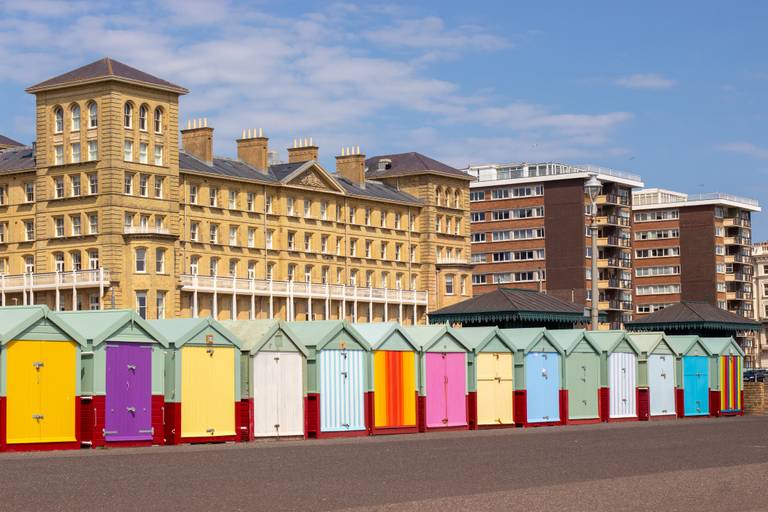 Colorful beach huts line a promenade with a historic hotel and modern apartments in the background under a blue sky.