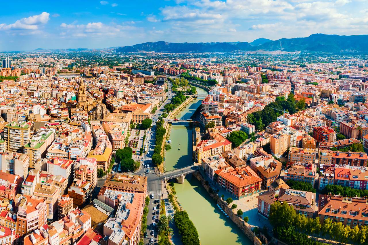 Vista aérea de Murcia atravesada por el río Segura, rodeada de edificios densamente poblados y montañas lejanas bajo un cielo parcialmente nublado.