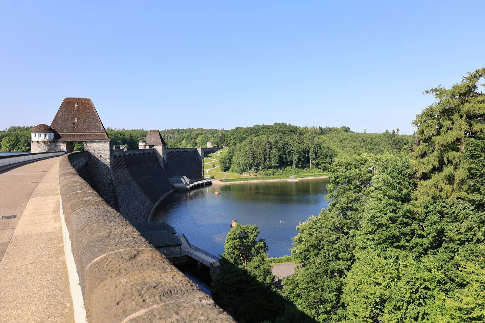 Ein Betondamm mit mehreren Türmen erstreckt sich über ein üppig grünes Tal und einen Stausee unter einem klaren blauen Himmel.