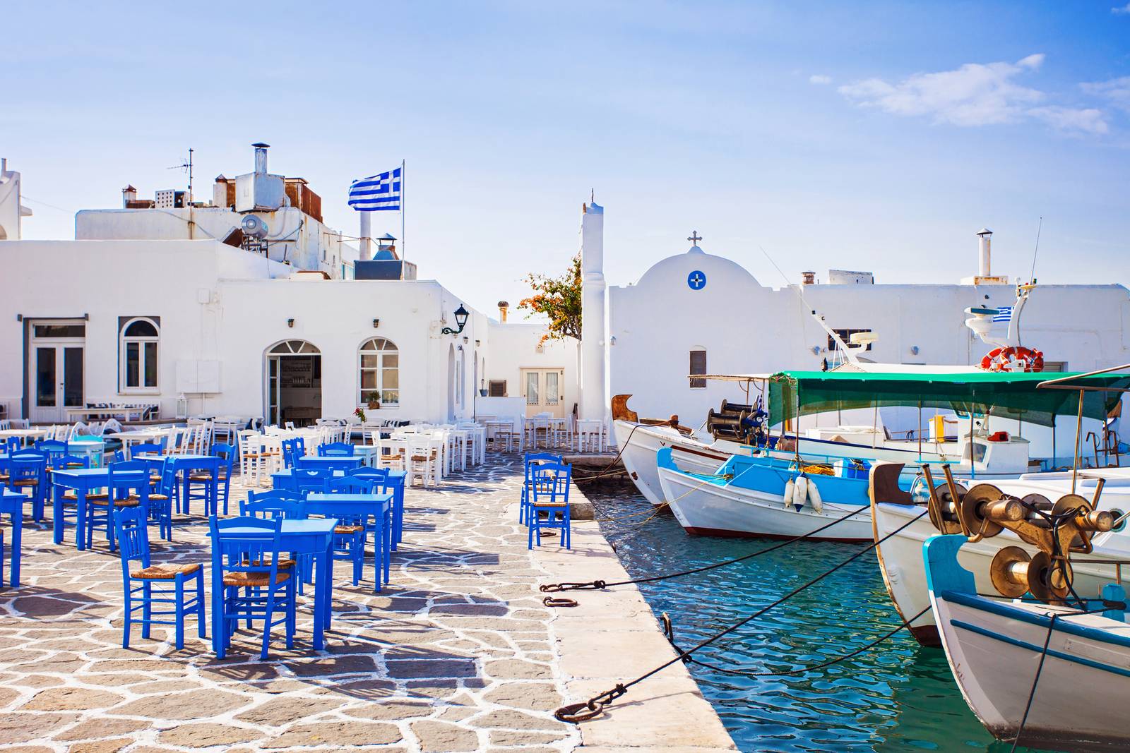 Greek seaside scene with white buildings, blue chairs, and tables on a stone patio. Boats are docked by the turquoise water under a clear sky.