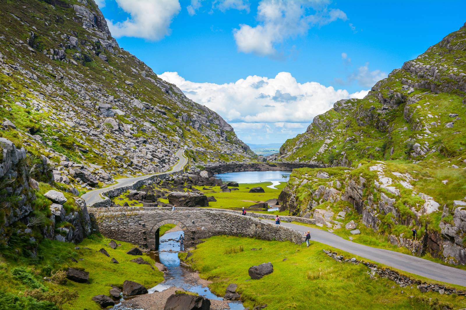 Scenic view of a winding road and stone bridge through lush green mountains with a small lake under a bright blue sky.