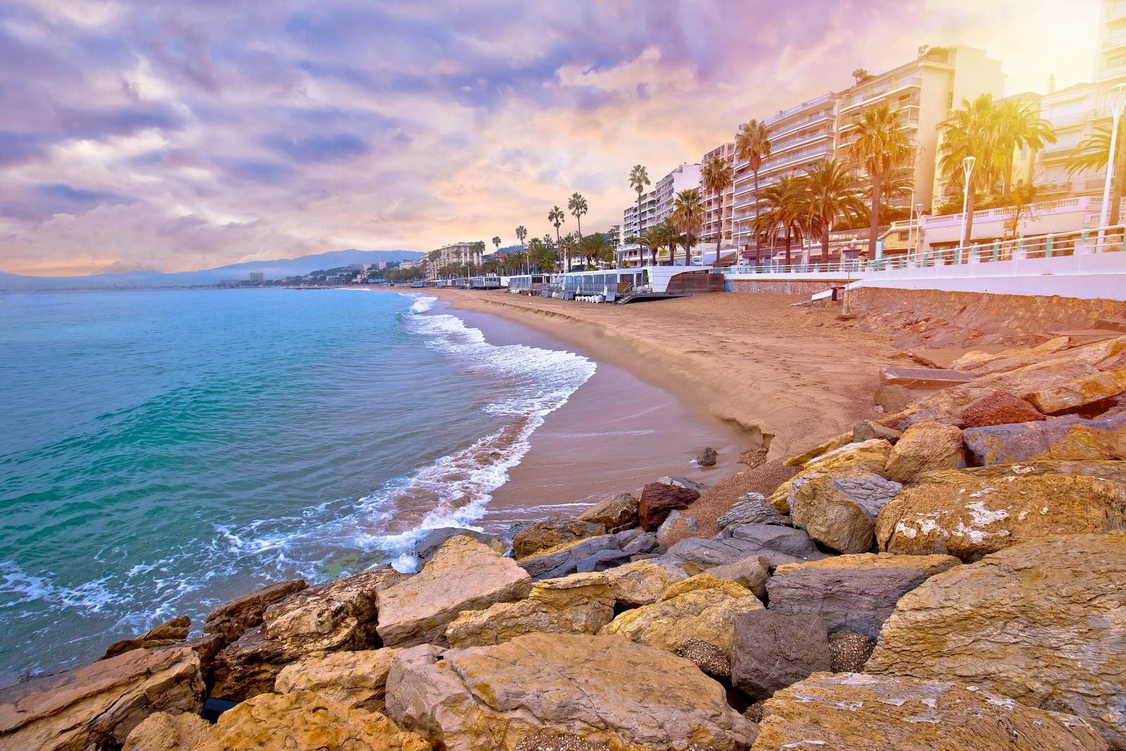 Beachfront with gentle waves, rocky foreground, palm trees, and tall buildings under a colorful sunset sky.