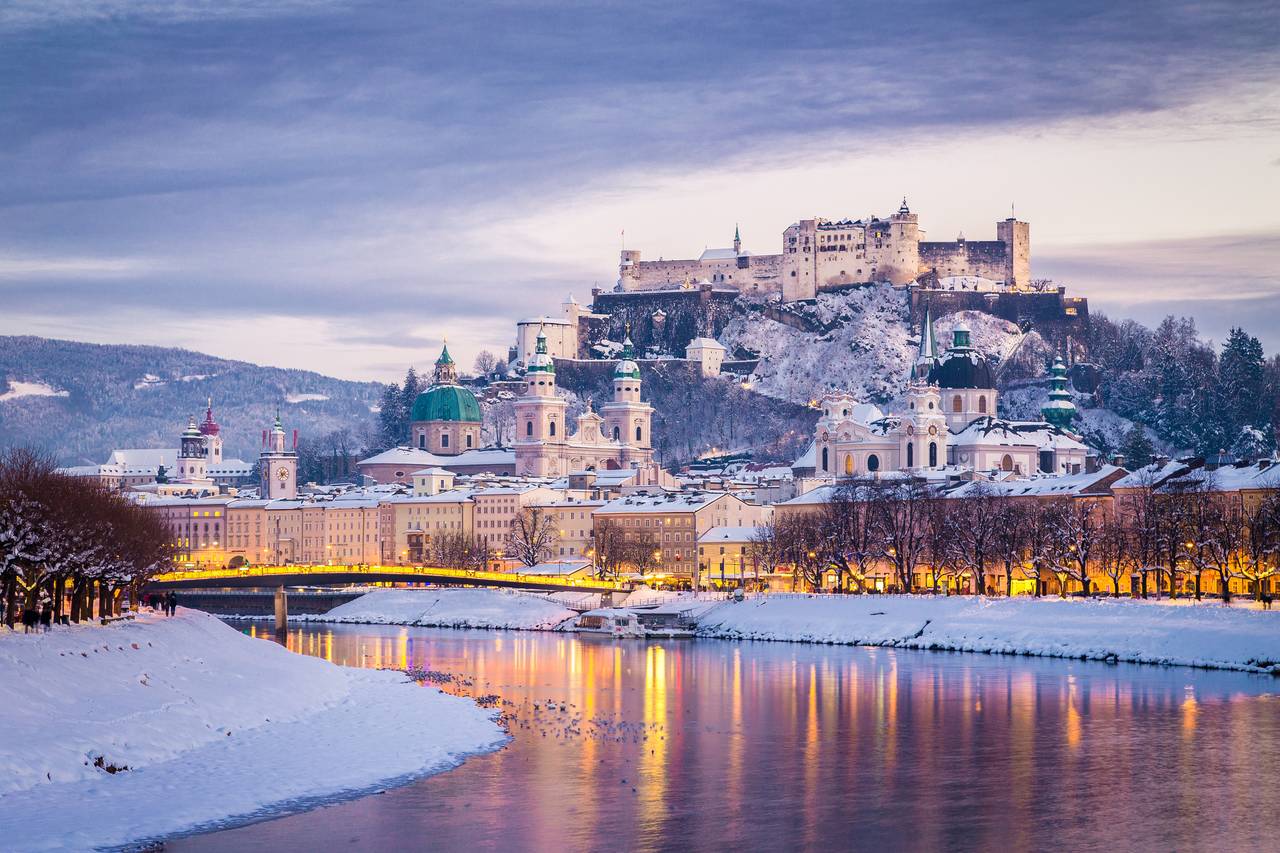 Verschneite Stadtsilhouette von Salzburg mit einer historischen Festung auf einem Hügel, die sich bei Einbruch der Dämmerung im ruhigen Fluss spiegelt. Warmes, gelbes Licht lässt die Szenerie stimmungsvoll erstrahlen.