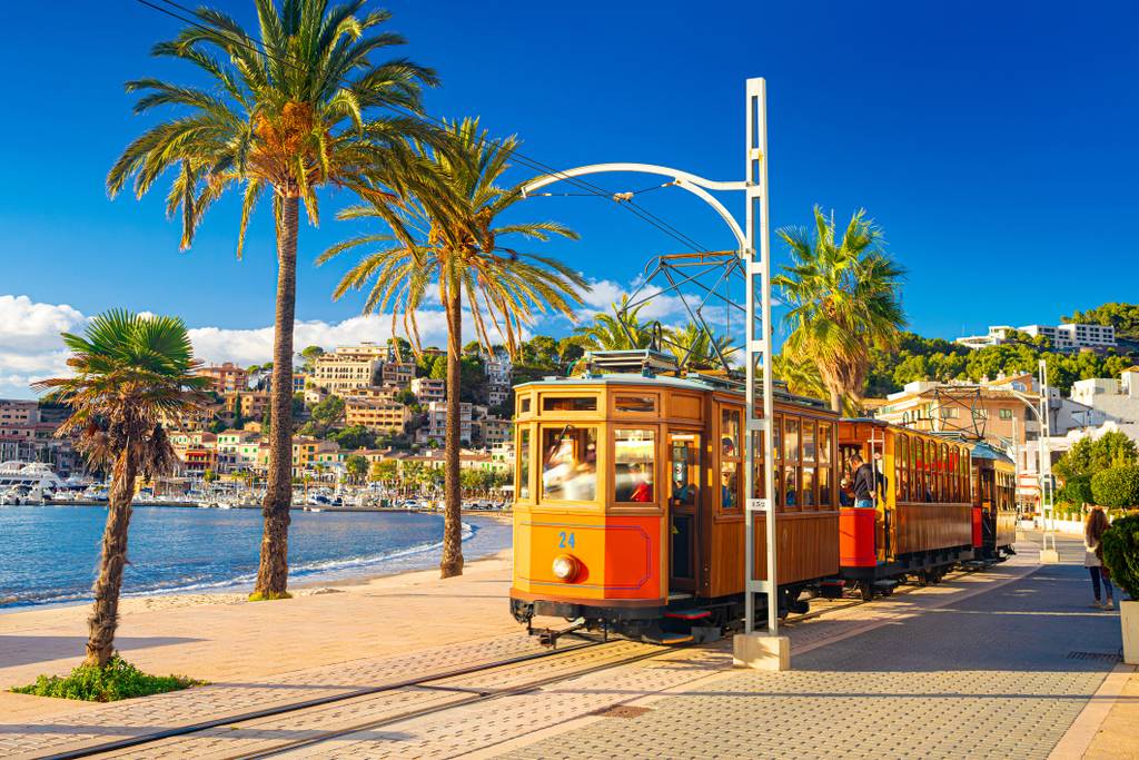 Tram d'epoca a Sóller accanto a un lungomare con palme, cielo soleggiato e un bel panorama urbano sullo sfondo