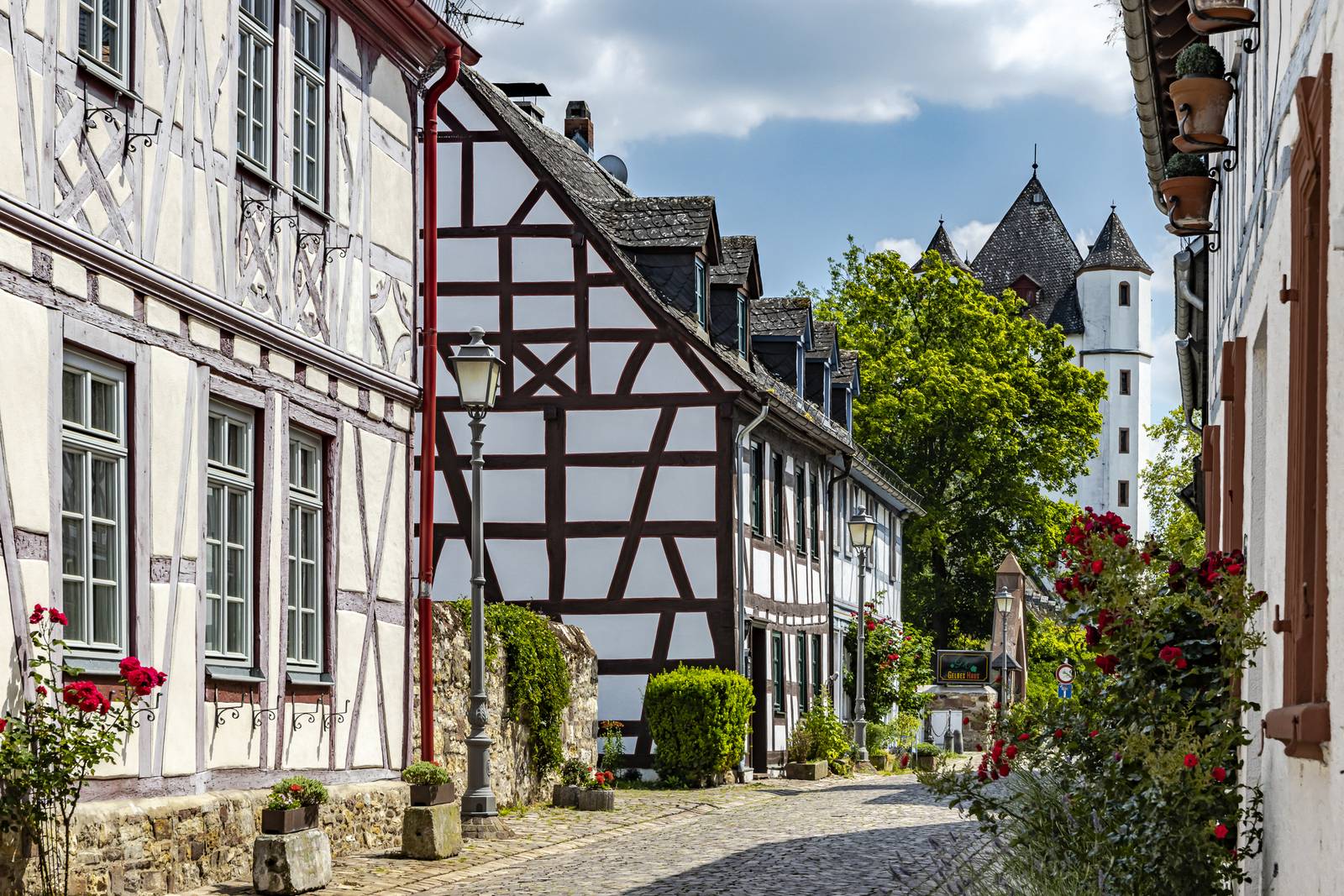 Kopfsteinpflasterstraße mit Fachwerkhäusern und blühenden Blumen, die zu einem Turm unter blauem Himmel mit Wolken führt