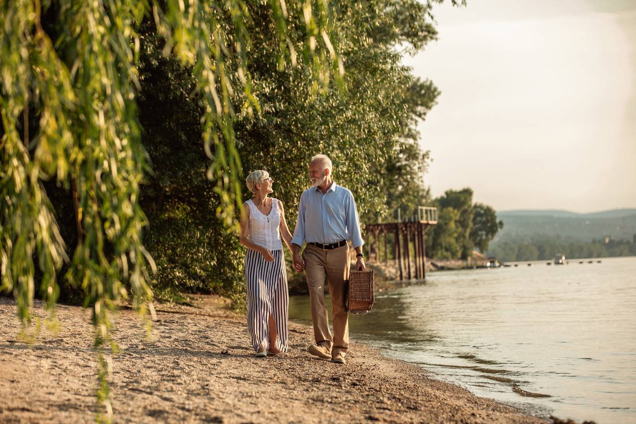 Ein älteres Paar spaziert Hand in Hand an einem See entlang, trägt einen Picknickkorb und ist von Bäumen unter einem klaren Himmel umgeben.