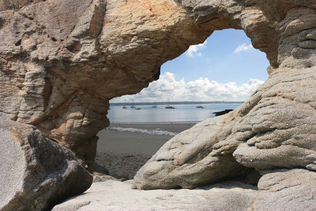 Une arche rocheuse naturelle encadre une plage paisible avec des voiliers naviguant sur une eau calme sous un ciel partiellement nuageux, idéale pour un séjour en chambre d'hôtes à Concarneau en Bretagne.