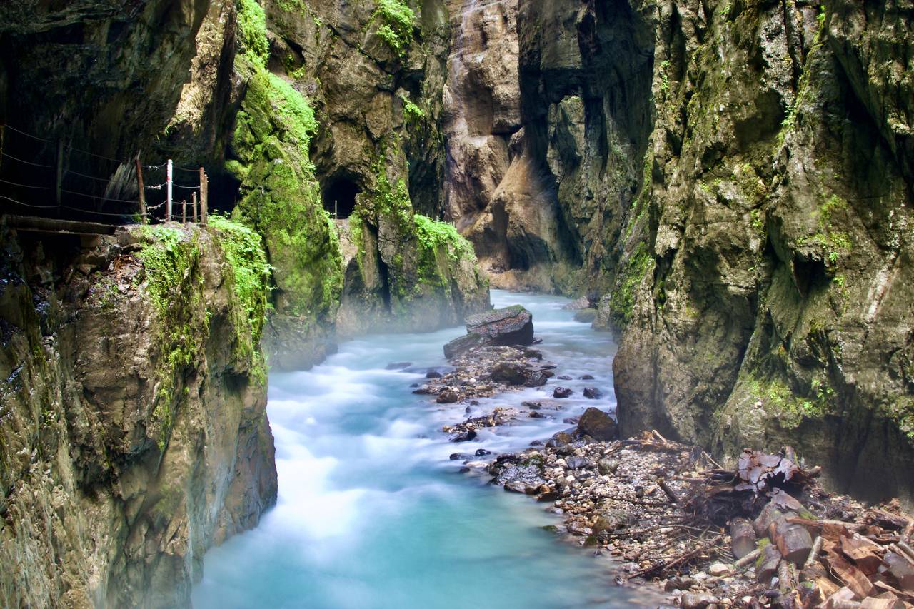 Eine schmale, felsige Schlucht mit türkisfarbenem Wasser, das schnell hindurchfließt, umgeben von steilen, moosbewachsenen Felswänden und einem schmalen Pfad auf der linken Seite.