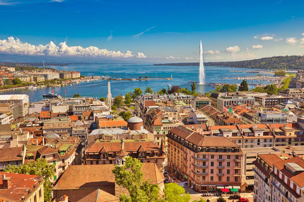 Aerial view of a cityscape with red-roofed buildings by a large lake, featuring a prominent water fountain and distant hills under a blue sky.