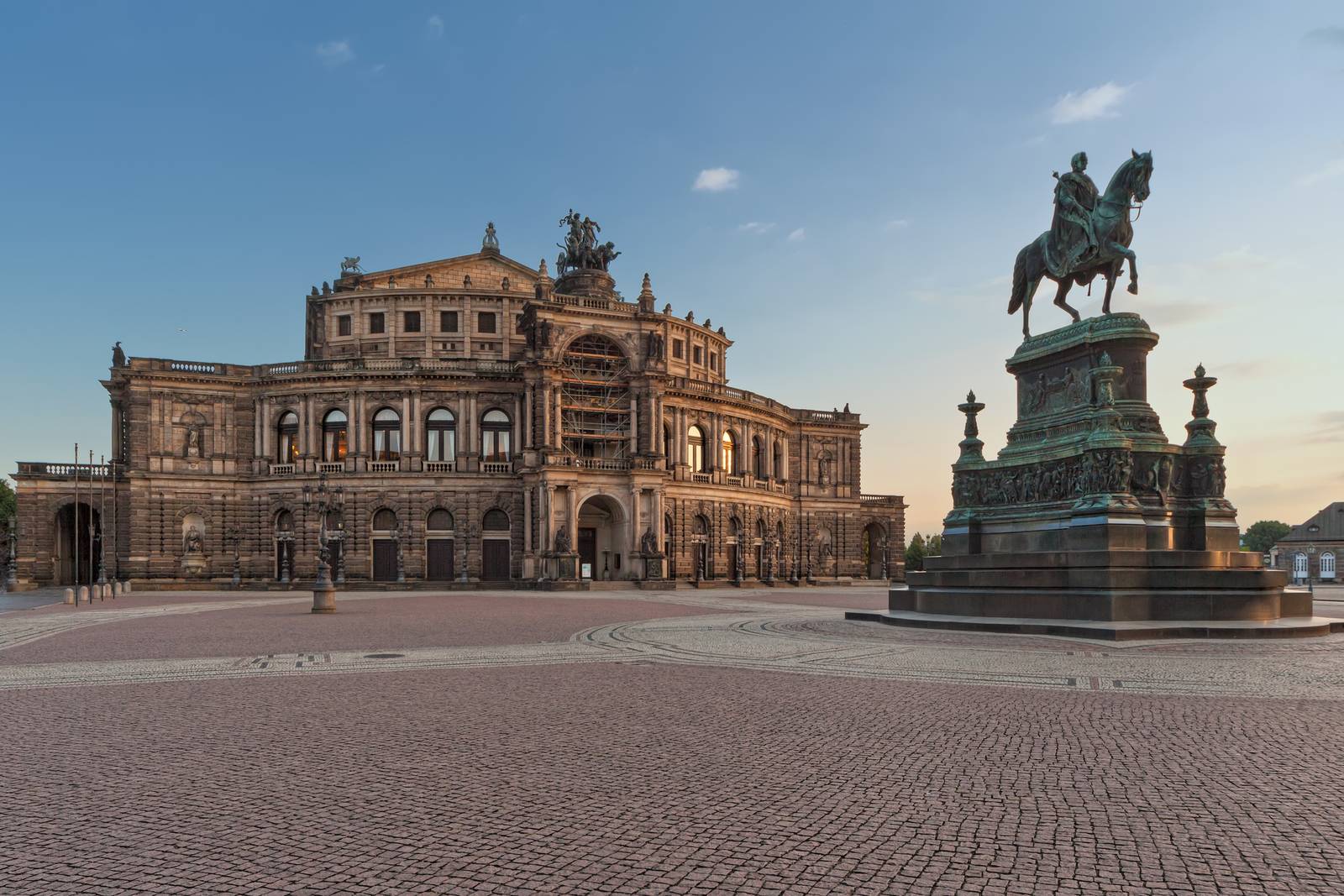 Historisches Opernhaus mit kunstvoller Architektur, einer großen Reiterstatue im Vordergrund und einem Kopfsteinpflasterplatz unter klarem Himmel