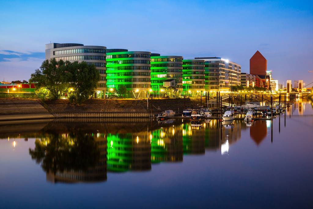 Moderne Gebäude am Flussufer mit grüner Beleuchtung, die sich im ruhigen Wasser spiegelt, Boote entlang des Ufers und ein Zwielichthimmel darüber.