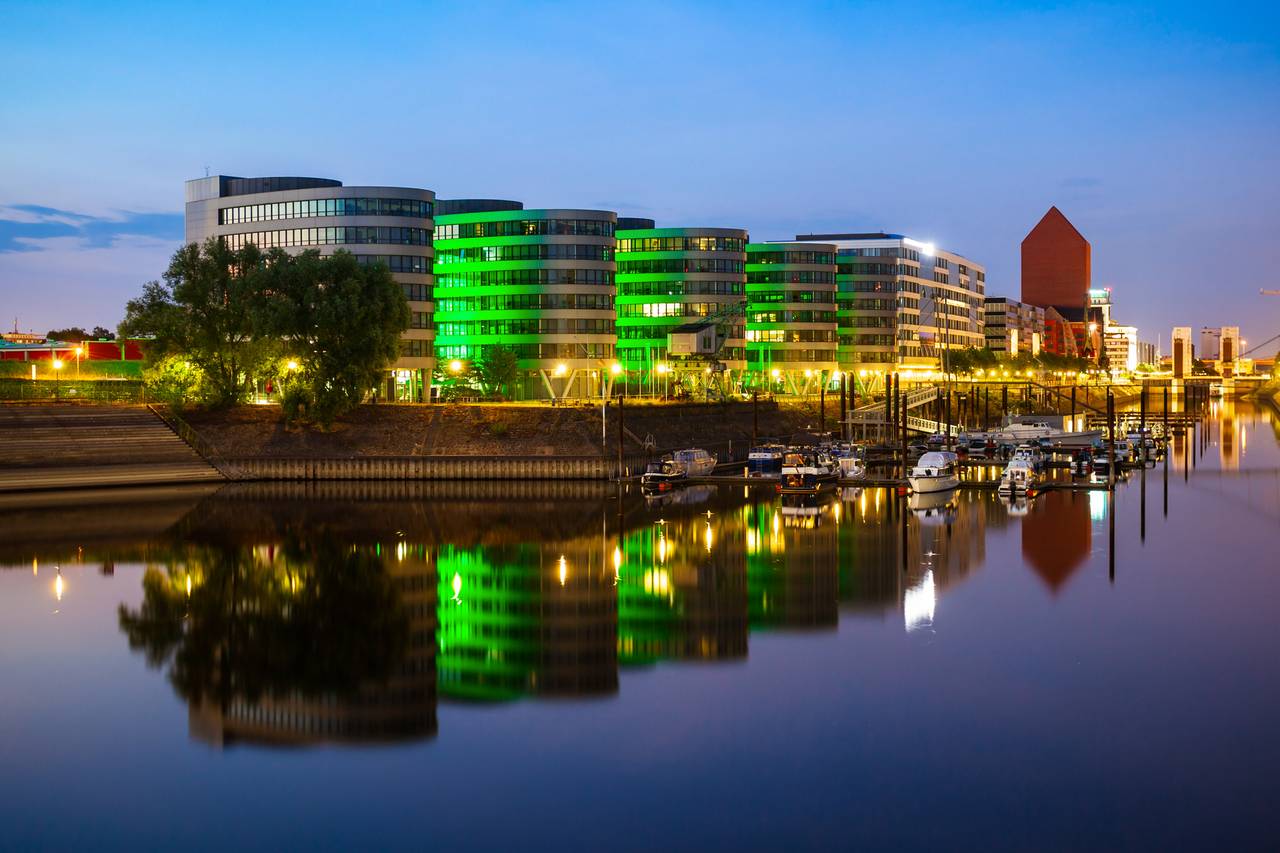 Moderne Gebäude am Flussufer mit grüner Beleuchtung, die sich im ruhigen Wasser spiegelt, Boote entlang des Ufers und ein Zwielichthimmel darüber.